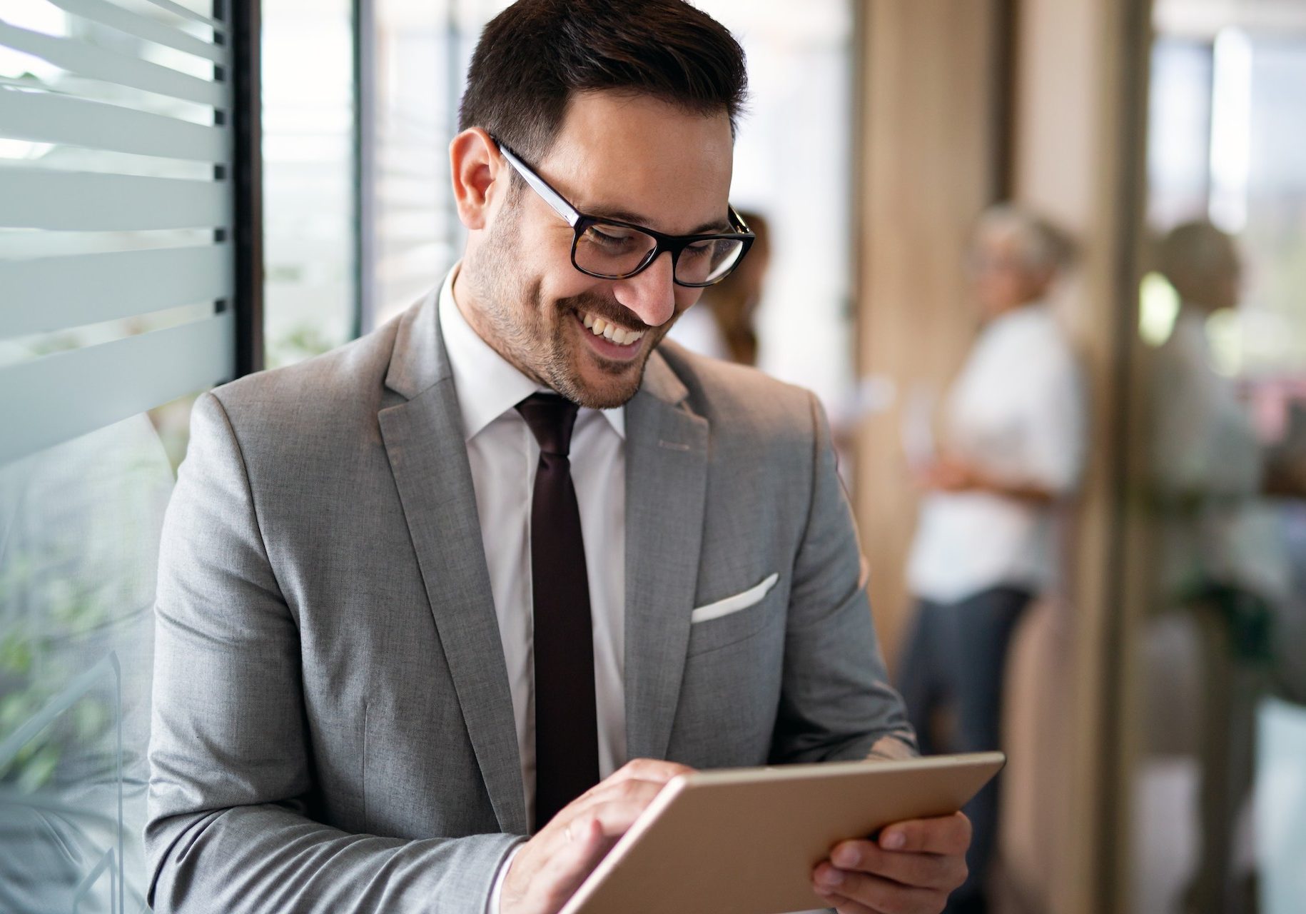 Happy young business man using a digital tablet while standing in front of windows in office