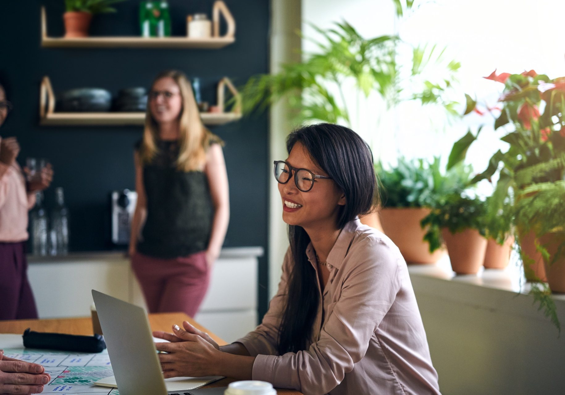 Smiling Asian businesswoman sitting at a table in a modern office using a laptop with colleagues working in the background